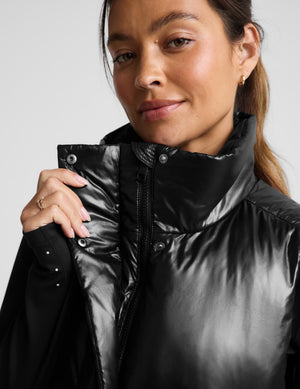 A woman modeling a black shiny quilted puffer vest over a black turtleneck, paired with black flared pants and white sneakers, against a light gray studio background.