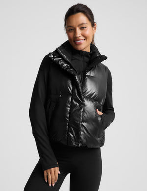 A woman modeling a black puffer vest with a shiny, quilted texture over a black long-sleeve top and black leggings, set against a solid light gray background.