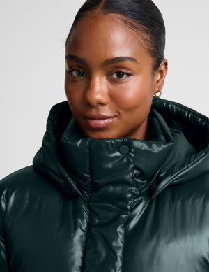 A close-up shot of a model wearing a shiny dark forest green puffer jacket with a high padded collar, set against a plain light gray background.