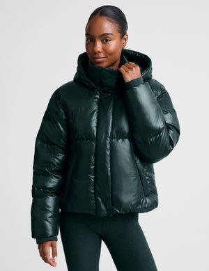 A woman wearing a shiny dark forest green puffer jacket with a high collar and hood, paired with green leggings, against a light gray studio background.