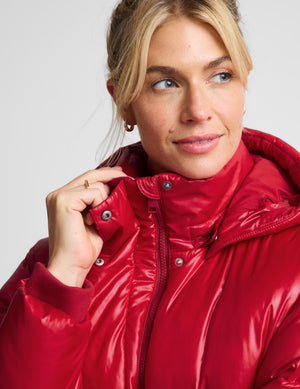A close-up of a woman wearing a shiny, high-collar red puffer jacket against a light gray studio background.