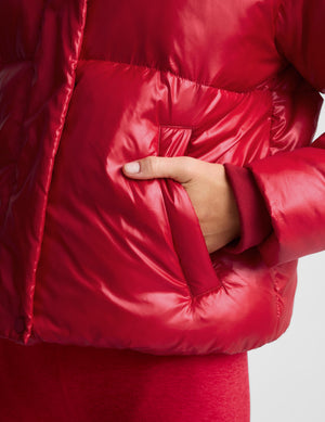 Close-up of a shiny red puffer jacket with a hand inserted into a front pocket, against a light gray background.