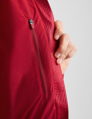 Close-up of a red jacket with a vertical zippered pocket and a hand pulling the zipper tab to demonstrate functionality, set against a light gray background.