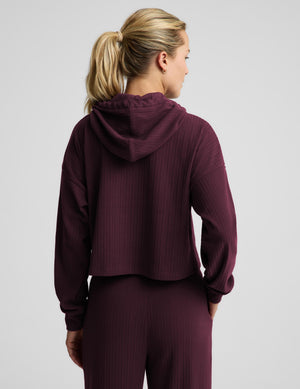 A woman modeling a matching ribbed lounge set in deep burgundy, featuring a midway length long-sleeve hoodie with drawstrings and coordinating drawstring pants, against a solid light gray background.