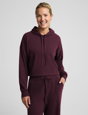 A woman modeling a matching ribbed lounge set in deep burgundy, featuring a midway length long-sleeve hoodie with drawstrings and coordinating drawstring pants, against a solid light gray background.