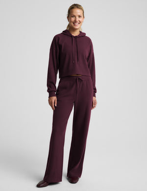 A woman modeling a matching ribbed lounge set in deep burgundy, featuring a midway length long-sleeve hoodie with drawstrings and coordinating drawstring pants, against a solid light gray background.