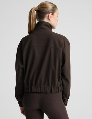 A woman shown from the back wearing a dark brown cropped zip-up jacket and matching dark brown leggings against a light gray studio background.