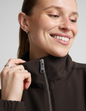 A woman modeling a dark brown zip-up jacket, matching dark brown cropped leggings, a white top, and black sneakers against a light gray studio background.