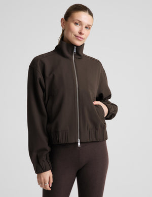 A woman modeling a dark brown zip-up jacket, matching dark brown cropped leggings, a white top, and black sneakers against a light gray studio background.