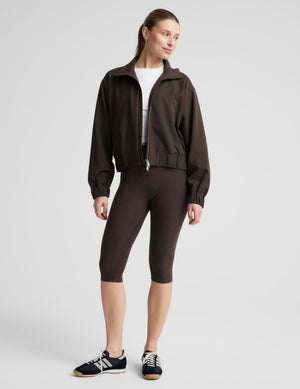 A woman modeling a dark brown zip-up jacket, matching dark brown cropped leggings, a white top, and black sneakers against a light gray studio background.