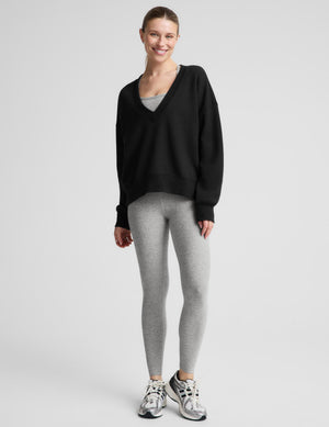 A woman modeling a black long-sleeve V-neck sweatshirt over a light gray tank top and heather gray leggings, photographed against a plain light gray studio background.
