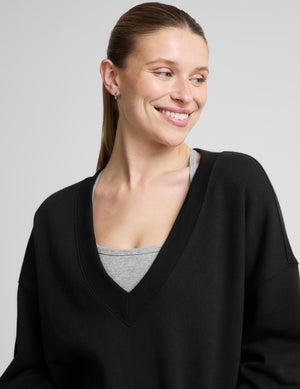 A woman modeling a black long-sleeve V-neck sweatshirt over a light gray tank top and heather gray leggings, photographed against a plain light gray studio background.