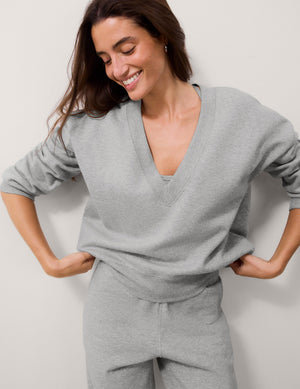 A woman wearing a light gray heathered V-neck long-sleeve top and matching jogger pants, posing with her hands on her hips against a plain light gray wall.