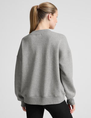 A woman shown from the back wearing a light gray crewneck sweatshirt and black pants, photographed against a plain light gray studio background.