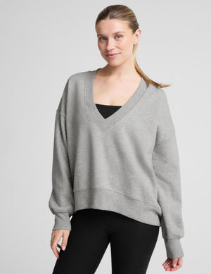 A woman wearing a light gray heathered v-neck sweatshirt with a relaxed fit, styled over a black top and black leggings, photographed against a neutral light gray studio background.