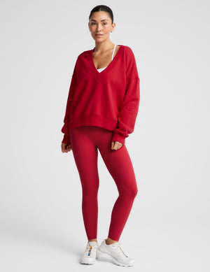 A woman modeling a matching red activewear set consisting of a V-neck sweatshirt and high-waisted leggings, paired with white sneakers, against a light gray studio background.