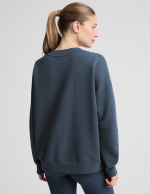 A woman with a blonde ponytail is shown from the back wearing an oversized crewneck sweatshirt in a dark slate blue color, paired with matching dark pants, against a light gray studio background.