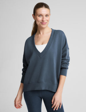 A woman wearing a slate blue v-neck sweatshirt over a white top and dark blue leggings, standing against a light gray studio background.