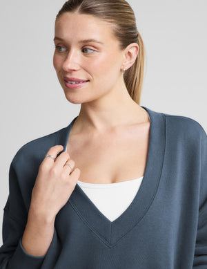 A woman wearing a slate blue-gray v-neck sweater over a white tank top, shown in a close-up shot against a neutral gray background.