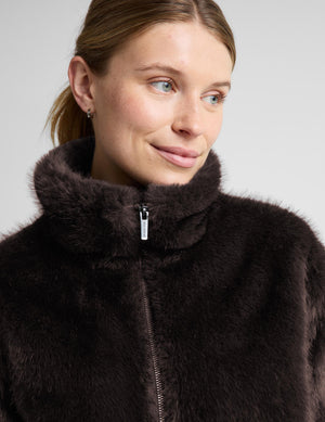 A woman modeling a dark brown faux fur jacket with a high collar and front zipper, paired with black pants, against a light gray studio background.