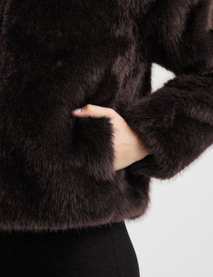 A woman modeling a dark brown faux fur jacket with a high collar and front zipper, paired with black pants, against a light gray studio background.