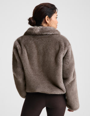 A woman shown from the back wearing a cropped, plush faux-fur jacket in taupe brown, paired with dark leggings, against a light gray studio background.