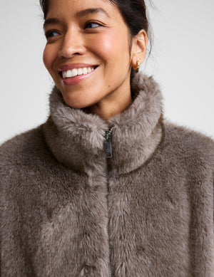 A close-up of a smiling woman wearing a plush, high-neck faux fur jacket in a taupe-brown color, set against a plain light gray background.
