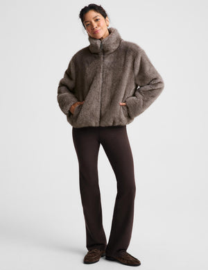 A model wearing a brown fuzzy faux-fur cropped jacket with a high mock neck, paired with dark brown pants and brown suede shoes, standing against a light gray studio background.