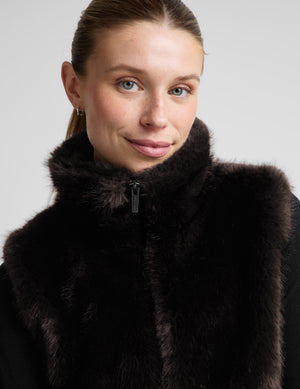 A close-up of a woman wearing a dark brown fur vest with a high collar and a black zipper, set against a plain light gray background.