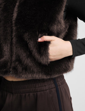 Close-up of a person wearing a dark brown faux fur cropped jacket over a black long-sleeve top and dark brown pants, with a hand pulling the fur to show texture.