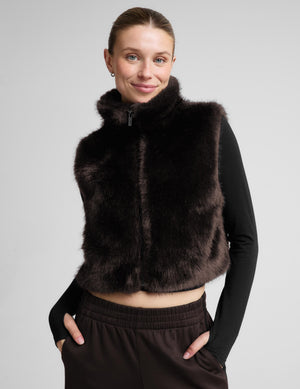 A woman wearing a dark brown faux fur vest over a black long-sleeve crop top and dark brown sweatpants, photographed against a neutral light gray studio background.