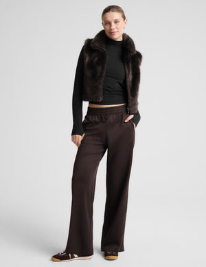 A woman modeling a dark brown faux fur vest over a black long-sleeve top and matching dark brown wide-leg trousers, standing against a light gray studio background.