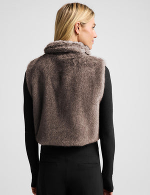 A woman modeling a cropped, high-collar faux fur vest in a taupe-brown color, worn over a black long-sleeve top and black leggings, against a light gray studio background.