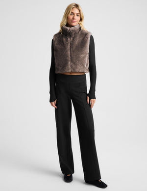 A woman modeling a cropped, high-collar faux fur vest in a taupe-brown color, worn over a black long-sleeve top and black leggings, against a light gray studio background.
