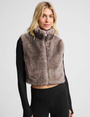 A woman modeling a cropped, high-collar faux fur vest in a taupe-brown color, worn over a black long-sleeve top and black leggings, against a light gray studio background.