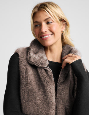 A woman modeling a cropped, high-collar faux fur vest in a taupe-brown color, worn over a black long-sleeve top and black leggings, against a light gray studio background.