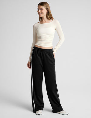 A woman modeling a fitted, long-sleeve crew-neck athletic top in cream, paired with black track pants, against a solid light gray background.