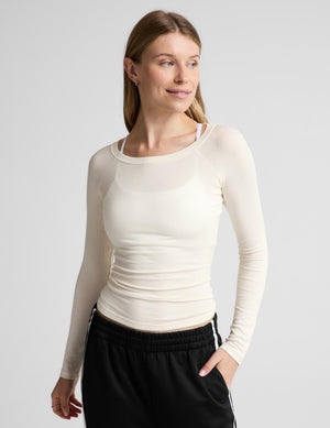 A woman modeling a fitted, long-sleeve crew-neck athletic top in cream, paired with black track pants, against a solid light gray background.