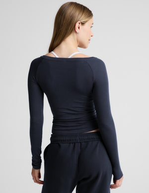 A woman modeling a dark navy long-sleeve athletic top with an off the shoulder neckline, paired with matching navy sweatpants, against a light gray studio background.