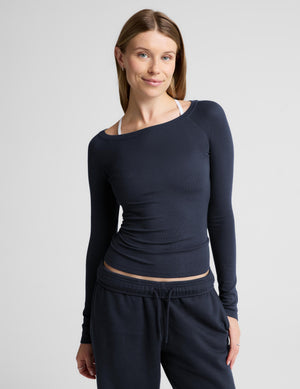 A woman modeling a dark navy long-sleeve athletic top with an off the shoulder neckline, paired with matching navy sweatpants, against a light gray studio background.