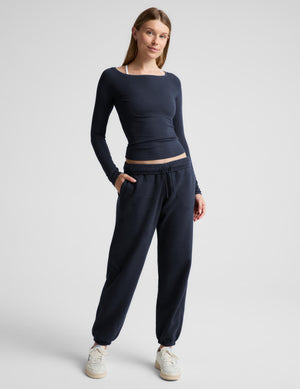A woman modeling a dark navy long-sleeve athletic top with an off the shoulder neckline, paired with matching navy sweatpants, against a light gray studio background.