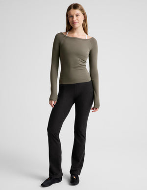 A woman modeling a fitted, long-sleeve ribbed top in olive green with a wide boat neckline, worn over a white camisole and paired with black leggings, against a light gray studio background.