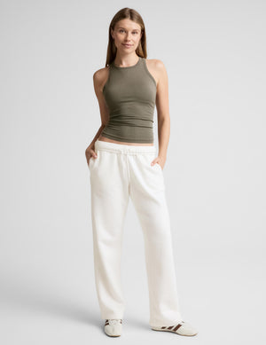A woman modeling a ribbed, sleeveless halter-style tank in olive green, paired with white sweatpants, against a solid light gray background.