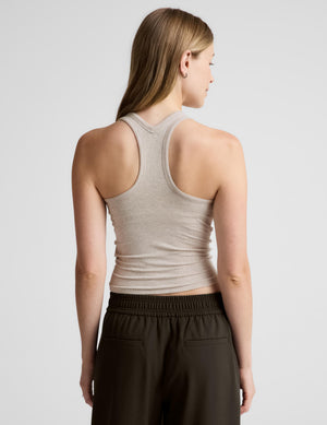 A woman shown from the back wearing a light beige racerback tank top and dark brown pants against a light gray studio background.