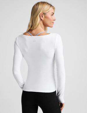 A blonde woman modeling a white long-sleeve scoop-neck top against a light gray studio background.