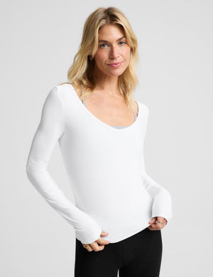 A blonde woman modeling a white long-sleeve scoop-neck top against a light gray studio background.