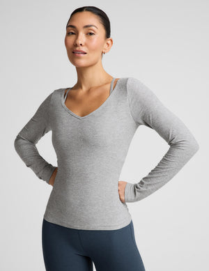A woman modeling a light heather gray long-sleeve V-neck athletic top, paired with dark blue leggings, against a solid light gray background.