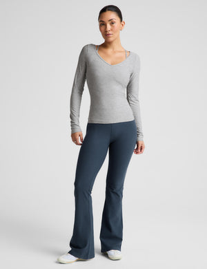 A woman modeling a light heather gray long-sleeve V-neck athletic top, paired with dark blue leggings, against a solid light gray background.
