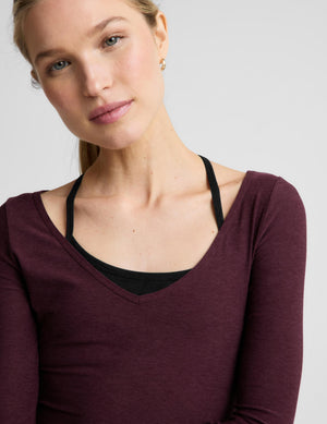 A woman modeling a long-sleeve scoop-neck athletic top in deep burgundy, worn over a black sports bra and paired with black leggings, against a light gray studio background.