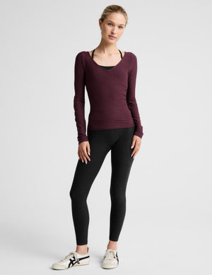 A woman modeling a long-sleeve scoop-neck athletic top in deep burgundy, worn over a black sports bra and paired with black leggings, against a light gray studio background.